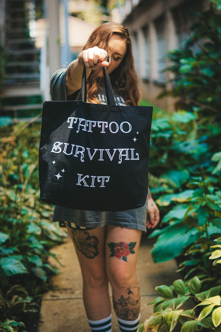 Woman with tattoos holding a Tote bag that says “tattoo survival kit”. A tote bag to pack the essentials to take to your tattoo appointment. 